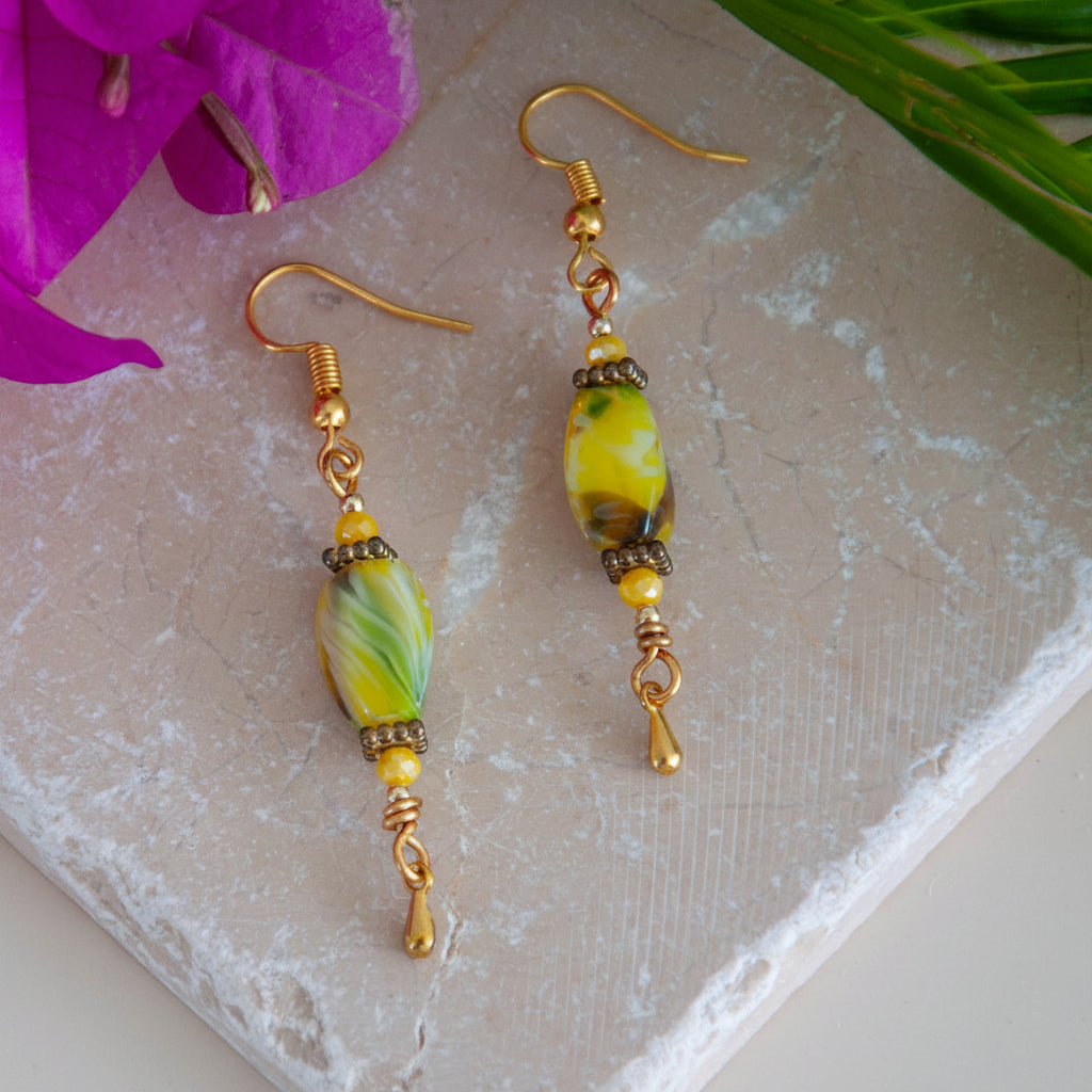 Handmade chartreuse twisted rectangle lampwork glass drop earrings with square gold-tone spacer beads, 3mm opaque yellow crystal, gold-tone teardrop dangle, and fishhook ear wires, laid on travertine tile with magenta orchid and tropical palm leaves.