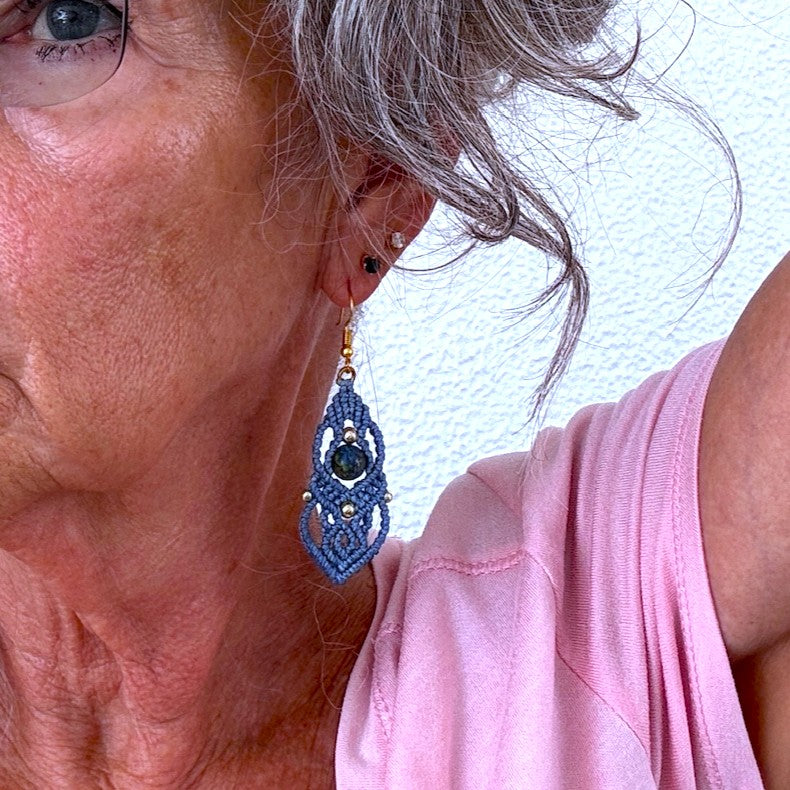 Handmade slate blue macrame drop earring with cascading lattice knotwork, 8mm round azurite gemstone center bead, 4mm and 3mm gold-tone metal beads, and gold-tone fishhook ear wire, shown worn on model with silver hair against white textured background.