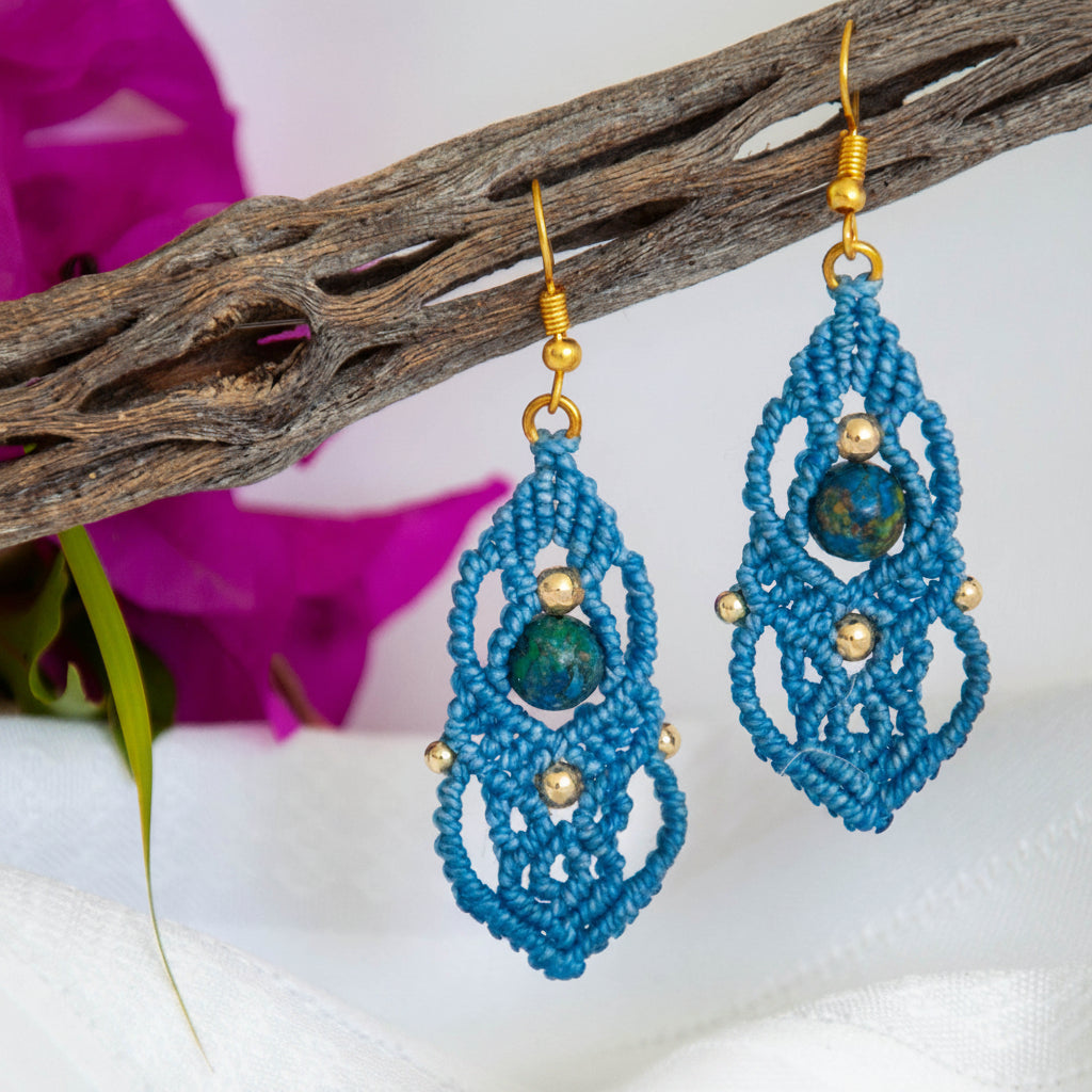 Pair of handmade slate blue macrame drop earrings with cascading lattice knotwork, 8mm round azurite gemstone center bead, 4mm and 3mm gold-tone metal beads, and gold-tone fishhook ear wires, hanging from rustic driftwood with magenta bougainvillea.