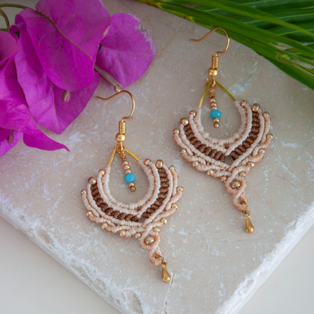 Handmade two-tone sand and chocolate brown macrame crescent chandelier earrings with galvanized gold seed beads, round metal bead accents, a faceted turquoise crystal rondelle center, gold-tone waterdrop charm, and gold-tone fish hook wires, laid on a travertine tile with magenta bougainvillea and tropical palm leaves.