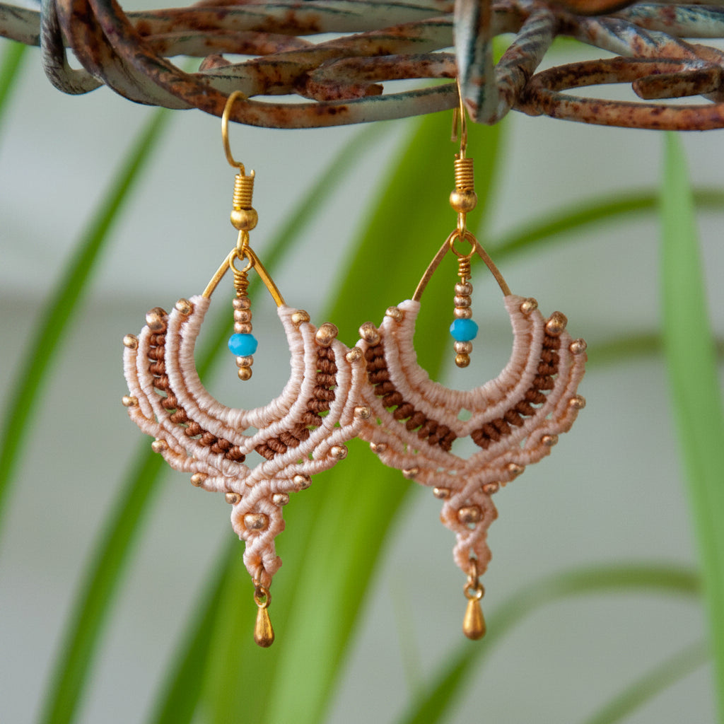Handmade two-tone sand and chocolate brown macrame crescent chandelier earrings with galvanized gold seed beads, round metal bead accents, a faceted turquoise crystal rondelle center, gold-tone waterdrop charm, and gold-tone fish hook wires, hanging from a rustic metal stand with tropical green leaves in the background.