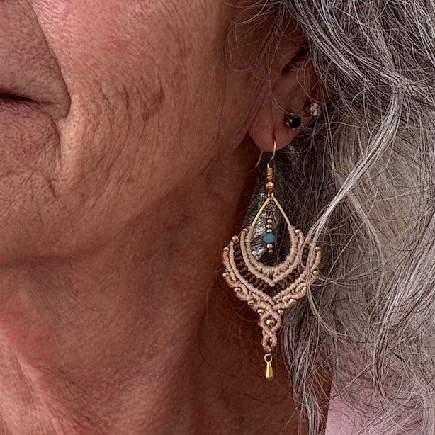 Handmade two-tone sand and chocolate brown macrame crescent chandelier earring with gold seed beads, turquoise crystal center, and gold-tone waterdrop charm, shown worn on model with silver hair.

