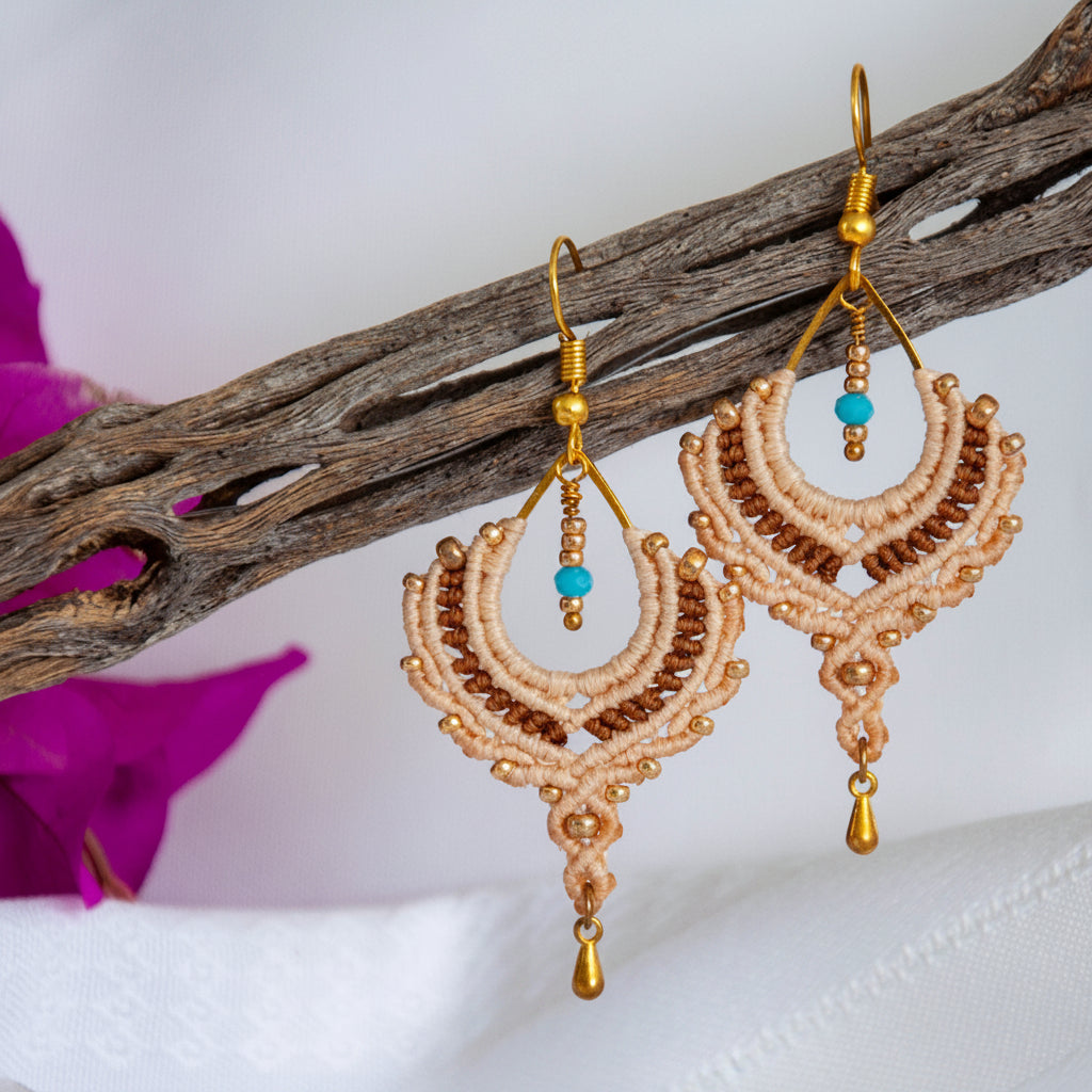 Handmade two-tone sand and chocolate brown macrame crescent chandelier earrings with galvanized gold seed beads, round metal bead accents, a faceted turquoise crystal rondelle center, gold-tone waterdrop charm, and gold-tone fish hook wires, hanging from a driftwood branch with magenta bougainvillea against a white background.