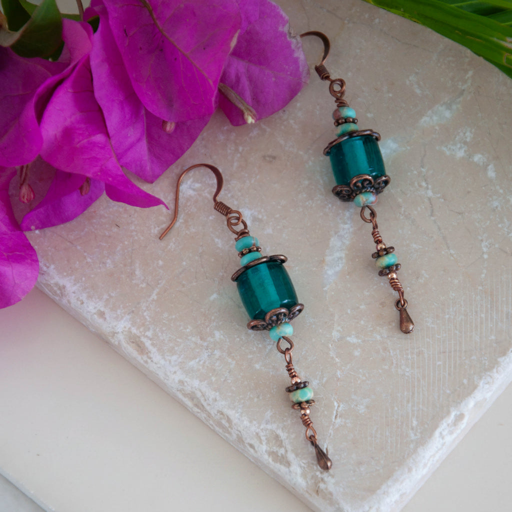 Handmade teal dyed glass drop earrings with copper filigree caps, abacus-shaped blue sea sediment beads, wire-wrapped copper waterdrop charms, and copper fish hook wires, laid flat on a natural stone surface with magenta bougainvillea flowers.