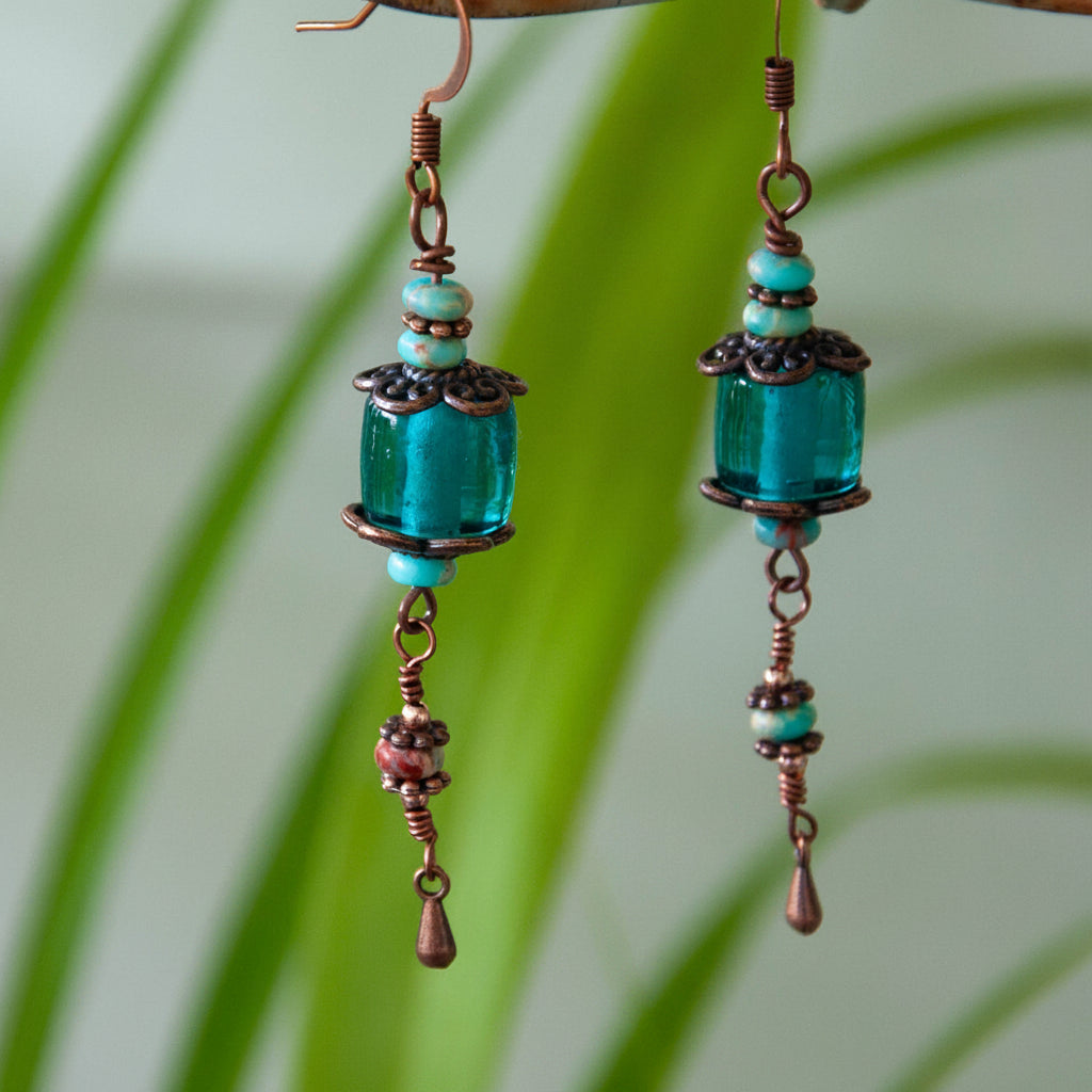 Handmade teal dyed glass drop earrings with copper filigree caps, abacus-shaped blue sea sediment beads, wire-wrapped copper waterdrop charms, and copper fish hook wires, hanging from a rustic branch with tropical green leaves.
