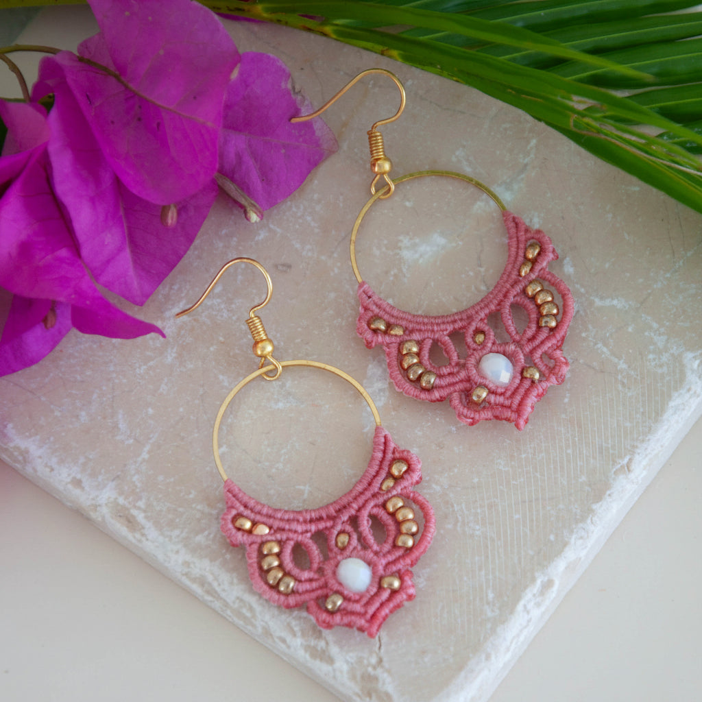 Handmade salmon pink macrame hoop drop earrings with a gold-tone hoop finding, galvanized gold seed beads, faceted iridescent white crystal rondelle center, and gold-tone fish hook wires, laid on a travertine tile with magenta bougainvillea flowers and tropical palm leaves.