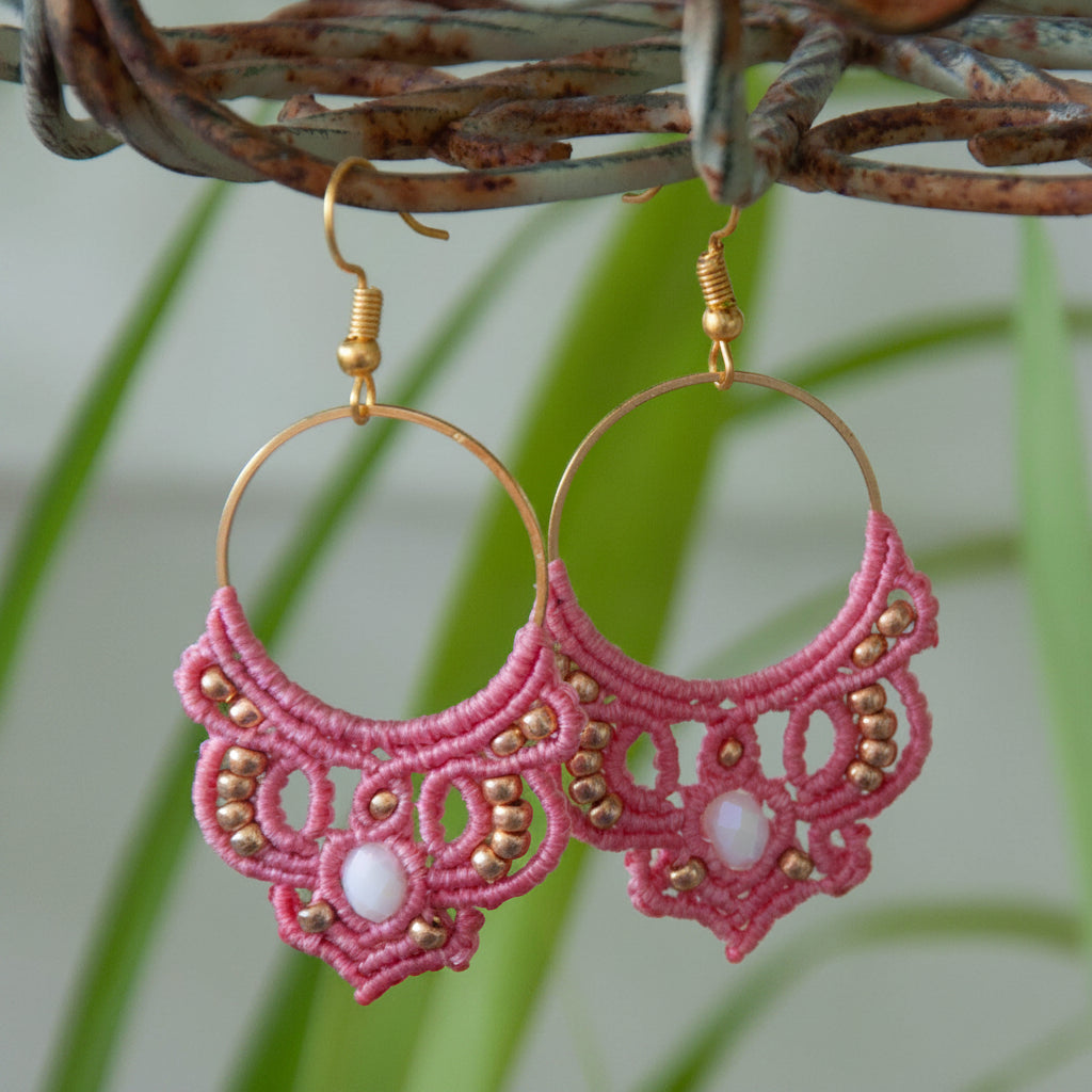 Handmade salmon pink macrame hoop drop earrings with a gold-tone hoop finding, galvanized gold seed beads, faceted iridescent white crystal rondelle center, and gold-tone fish hook wires, hanging from a rustic metal stand with tropical green leaves in the background.