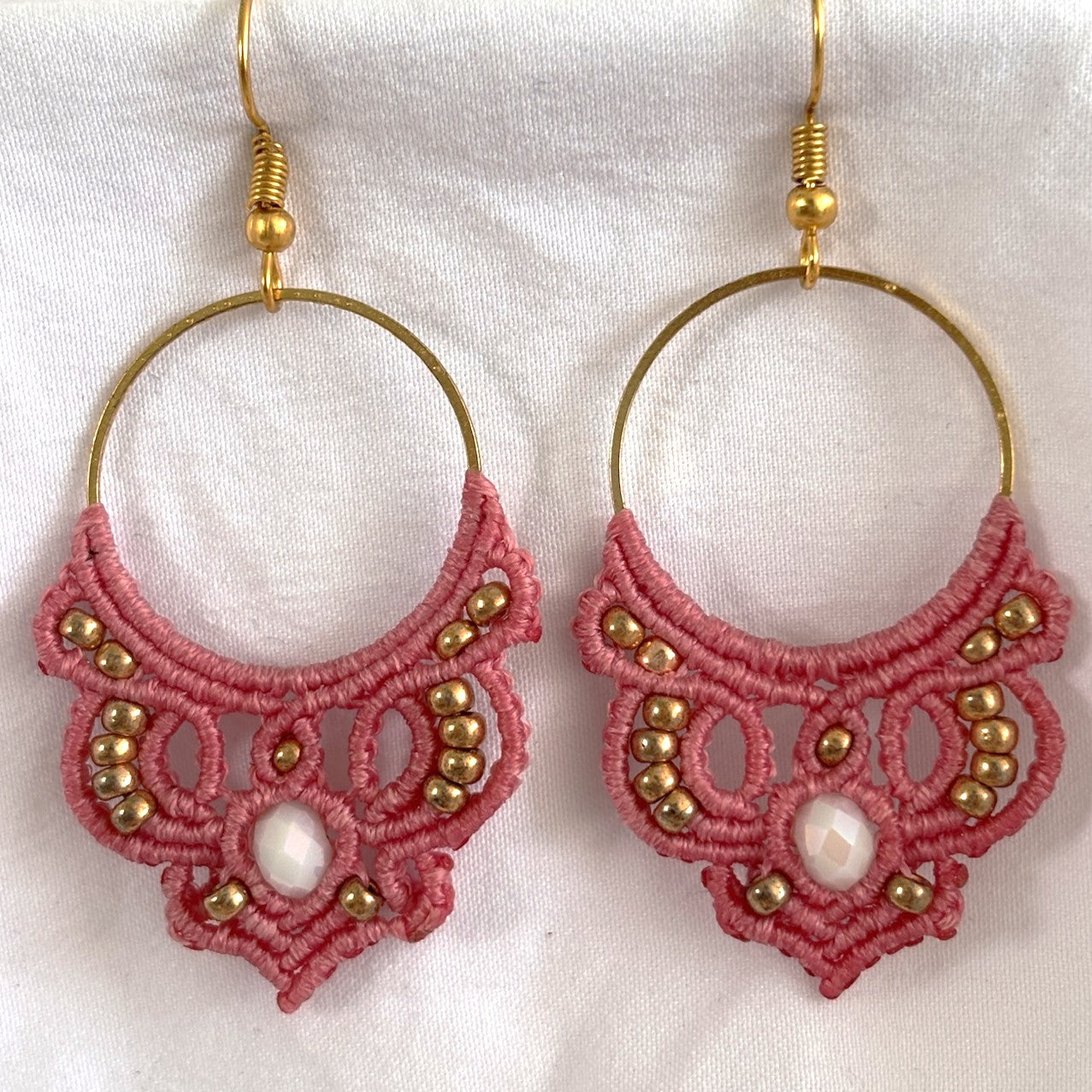 Pair of handmade salmon pink macrame hoop drop earrings with gold-tone hoop findings, galvanized gold seed beads, and faceted iridescent white crystal rondelle centers, displayed on white fabric background.