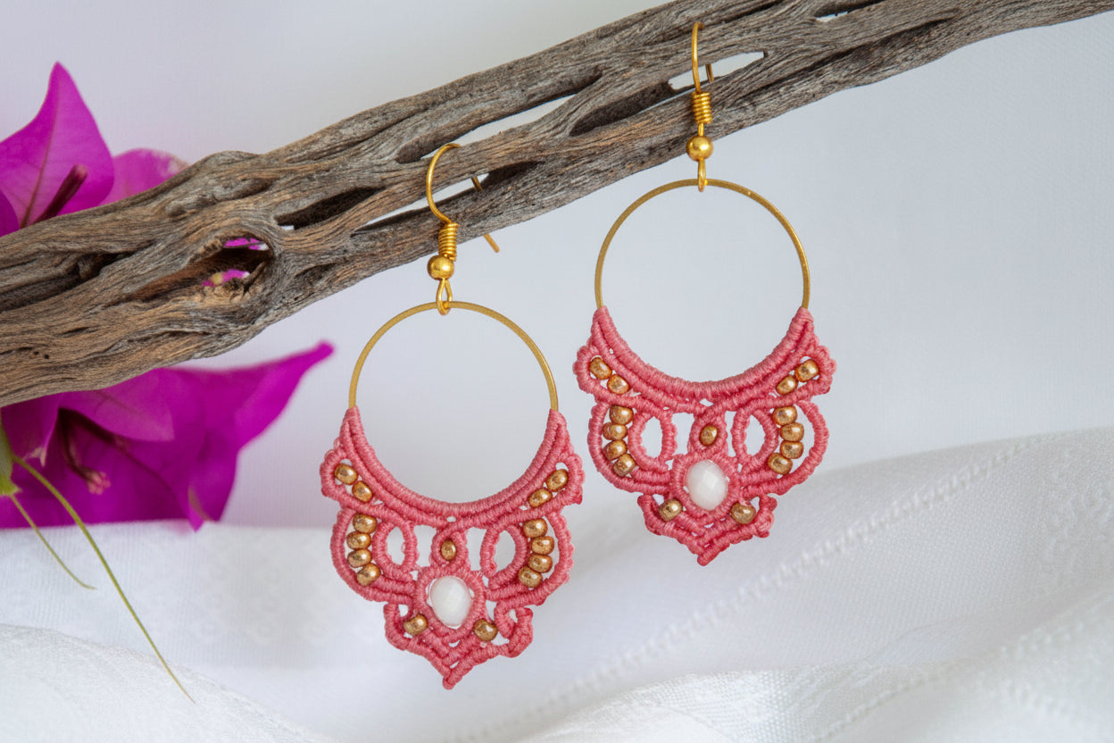 Handmade salmon pink macrame hoop drop earrings with a gold-tone hoop finding, galvanized gold seed beads, faceted iridescent white crystal rondelle center, and gold-tone fish hook wires, hanging from a driftwood branch with magenta bougainvillea flowers against a white background.
