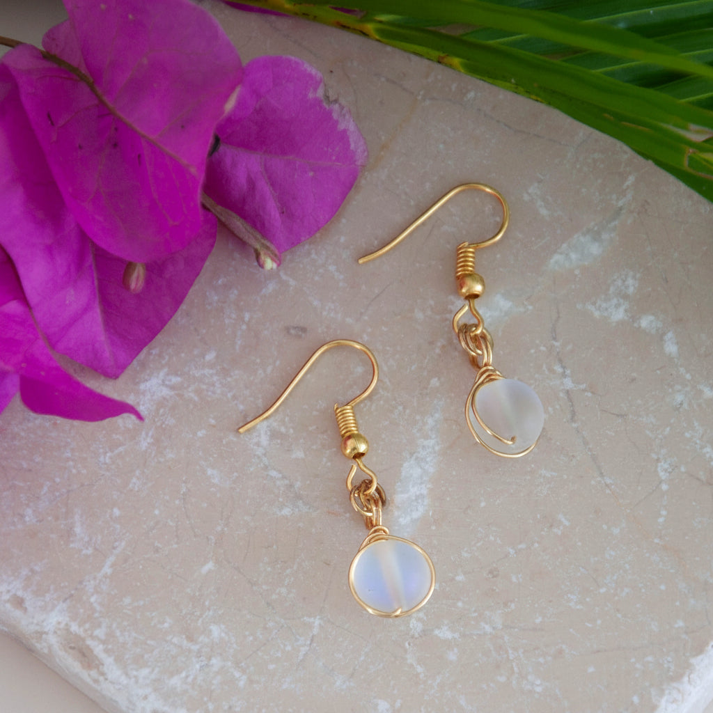 Handmade frosted clear mermaid glass drop earrings with brass wire wrapping and gold-tone fishhook ear wires, laid on travertine tile with magenta bougainvillea and tropical palm leaves.