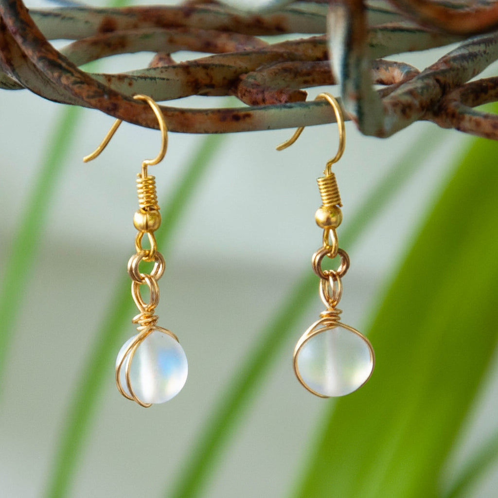 Handmade frosted clear mermaid glass drop earrings with brass wire wrapping and gold-tone fishhook ear wires, hanging from a rustic wire branch with tropical green leaves.