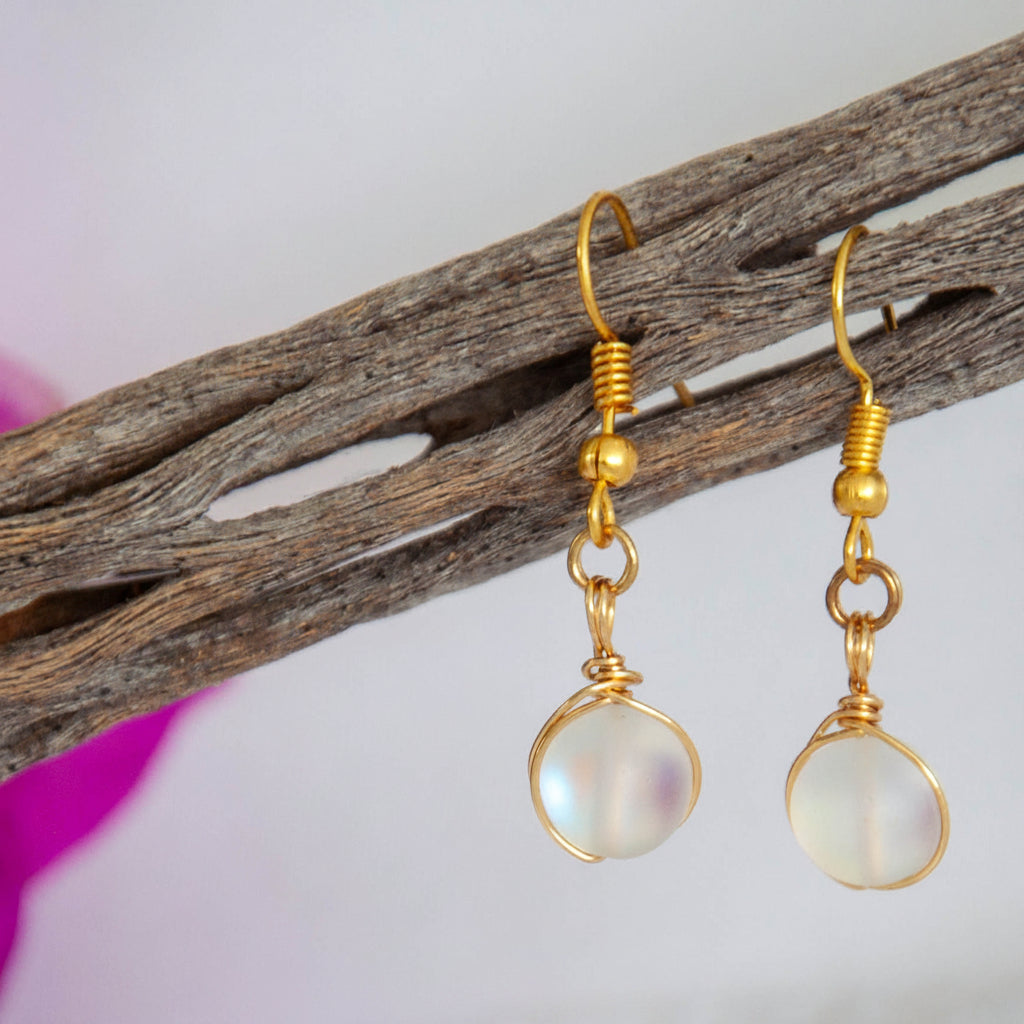 Handmade frosted clear mermaid glass drop earrings with brass wire wrapping and gold-tone fishhook ear wires, hanging from driftwood with a magenta flower.