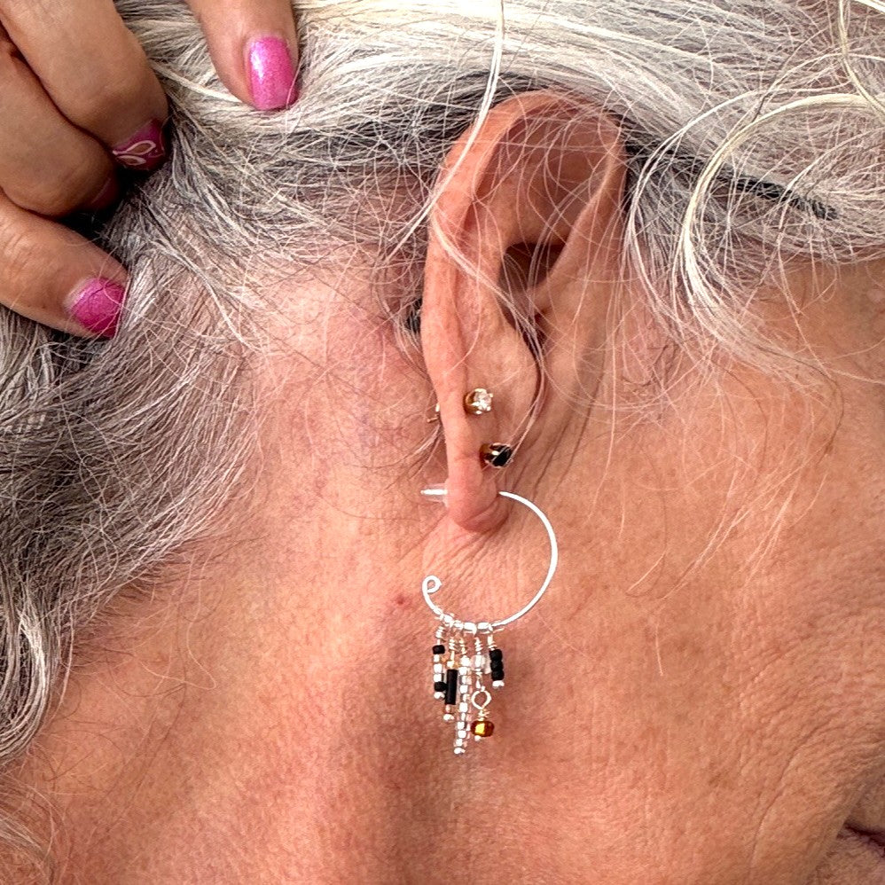 One silver-tone hoop earring with five wire-wrapped dangle strands of black bugle beads, clear seed beads, frosted white disc beads, and an amber faceted cube bead shown worn at the ear for scale