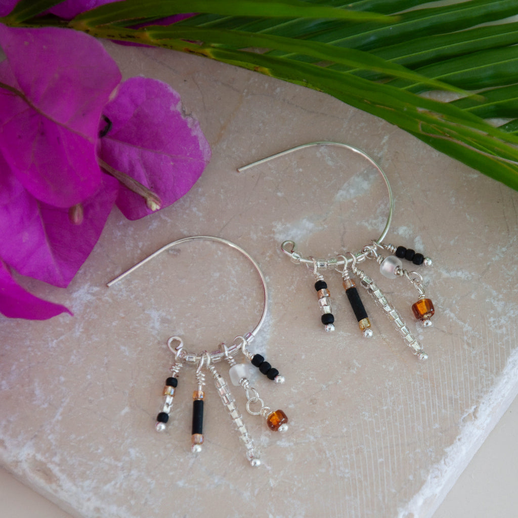 A pair of handcrafted silver-tone hoop earrings just under one inch in diameter with five wire-wrapped dangle strands of matte black bugle beads, clear seed beads, frosted white disc beads, and an amber faceted cube bead drop, displayed flat on a pink stone surface with a vibrant magenta bougainvillea flower and tropical palm fronds in the background.