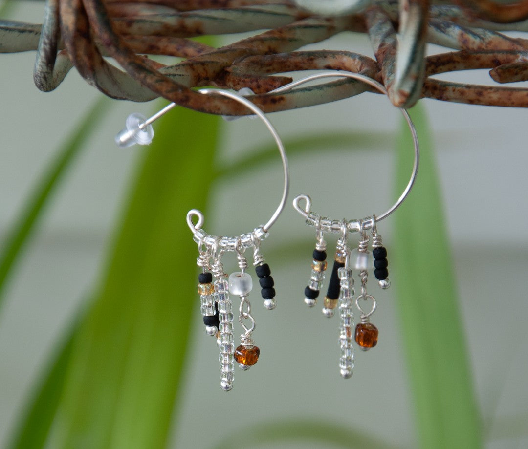 A pair of handcrafted silver-tone hoop earrings just under one inch in diameter with five wire-wrapped dangle strands of matte black bugle beads, clear seed beads, frosted white disc beads, and an amber faceted cube bead drop, hanging from a rustic twisted vine with soft-focus green tropical foliage in the background.