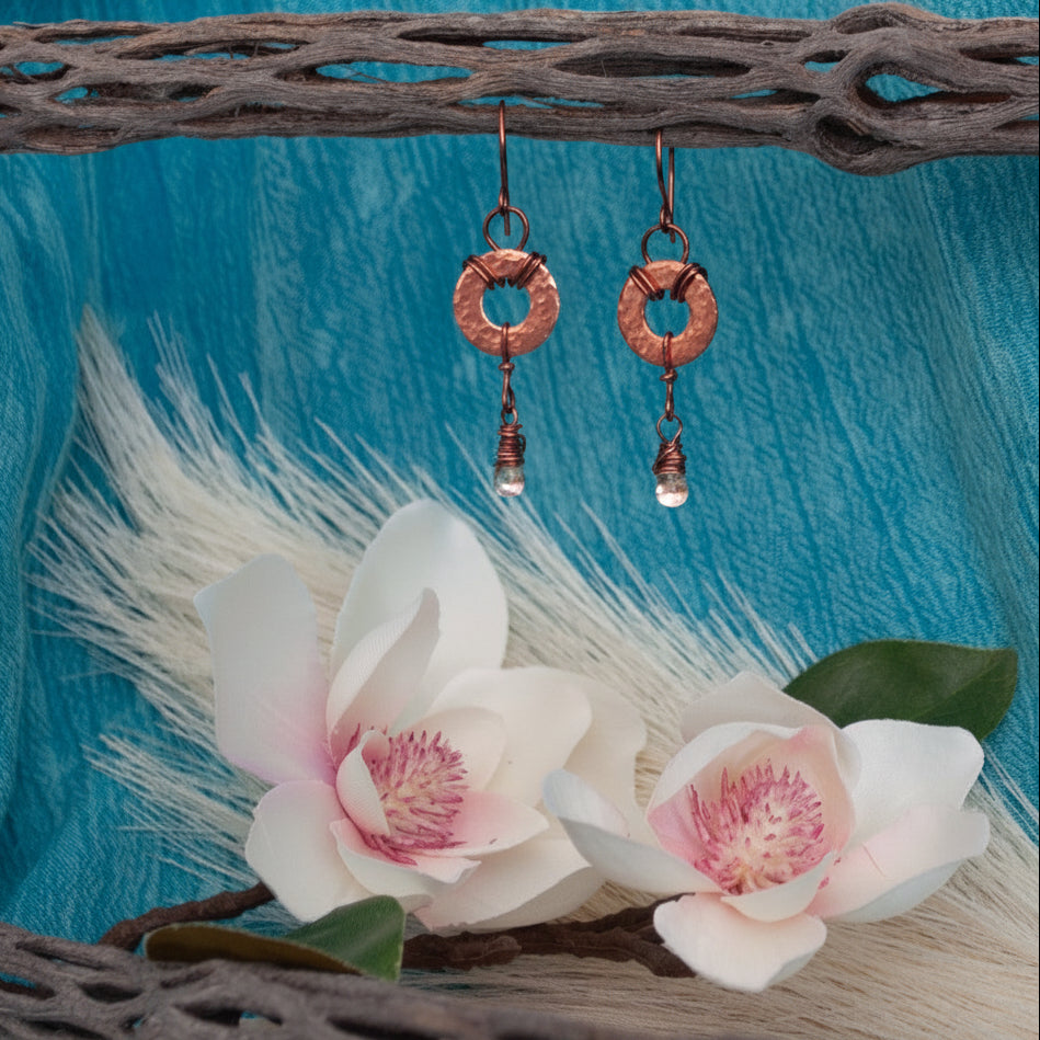 A pair of copper ring dangle earrings with small waterdrop beads, displayed on a rustic branch against a bright blue fabric backdrop with white magnolia flowers.