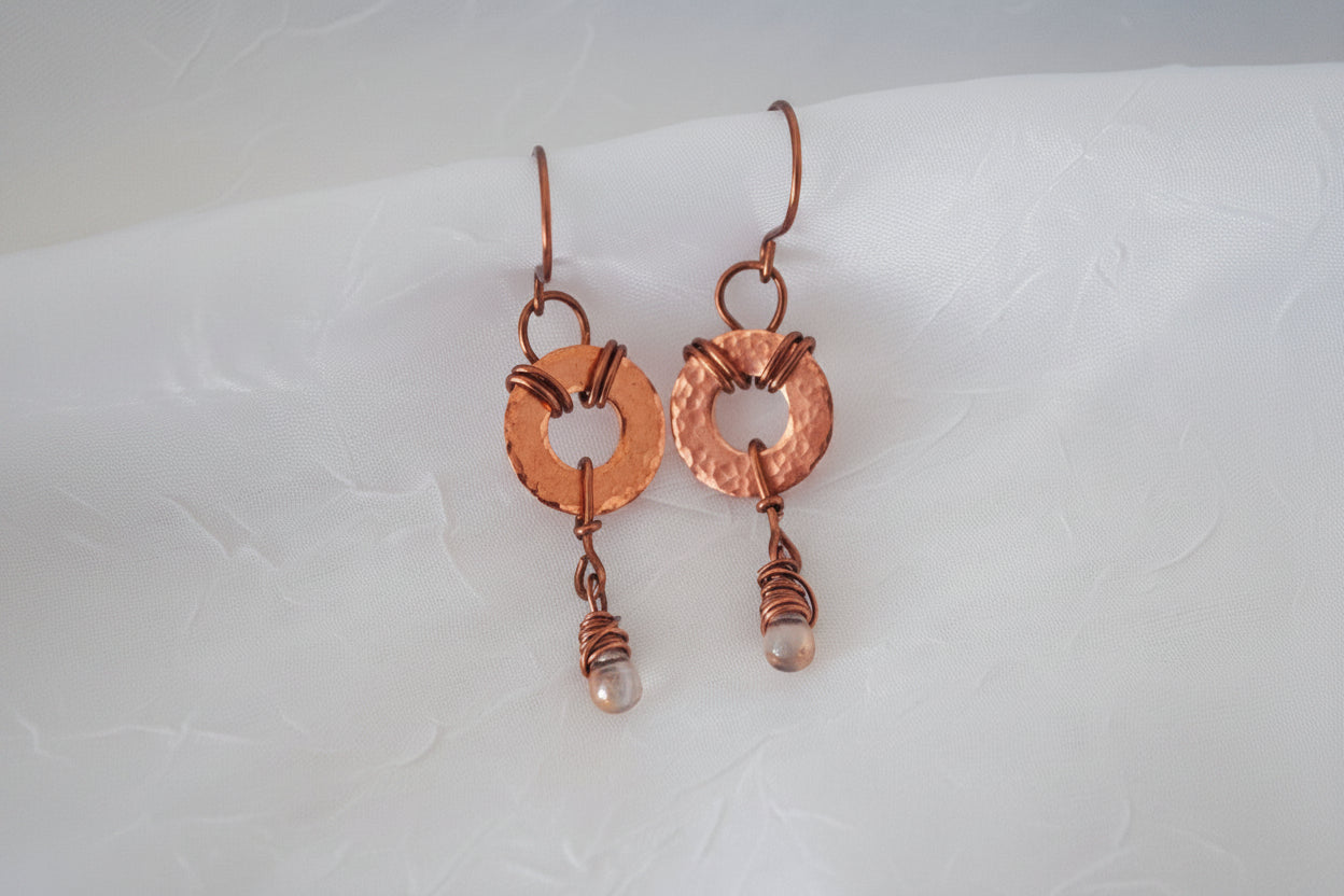 A pair of handmade copper and wire-wrapped drop earrings with small waterdrop beads, displayed on a textured white fabric background.
