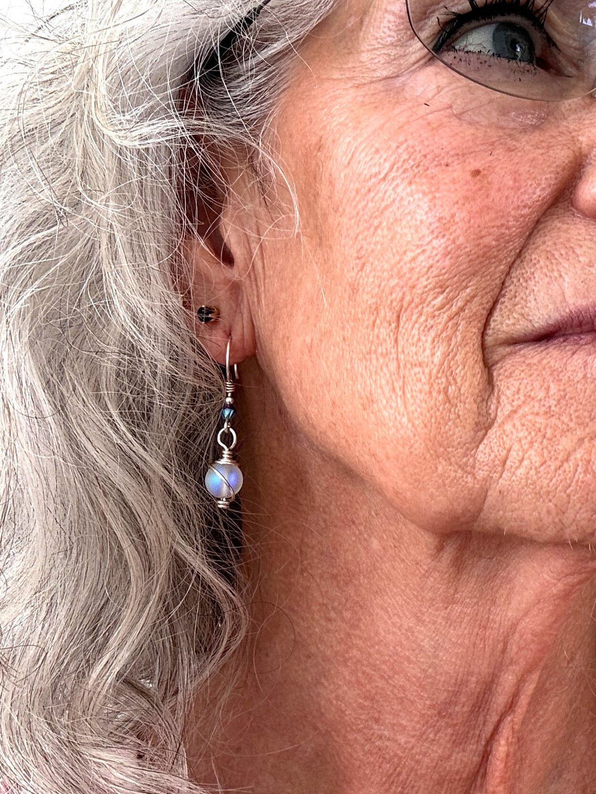 Mermaid glass earring with cosmic wire-wrapped drop and purple Swarovski bicone shown worn on model with silver hair, close-up showing blue iridescent flash in mermaid glass bead