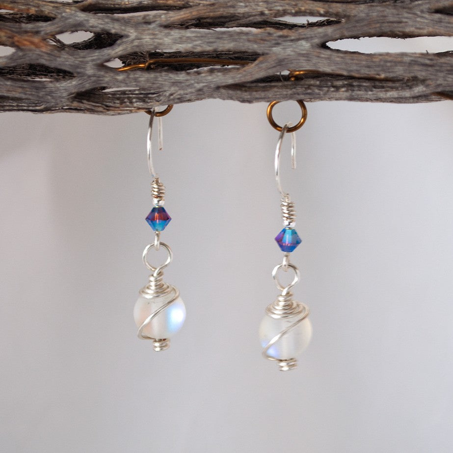 Handmade mermaid glass earrings with 8mm matte clear iridescent mermaid glass bead in cosmic wire wrap and hammered ear wires accented with purple Swarovski bicone, displayed on driftwood with white background