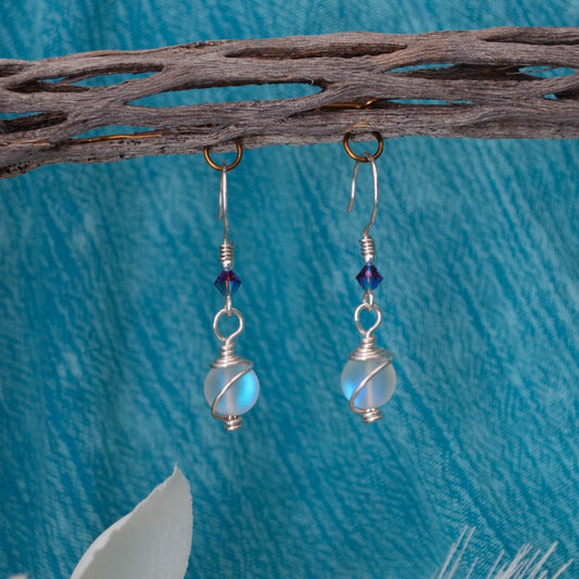 Mermaid glass earrings with cosmic wire-wrapped drop and purple Swarovski bicone hanging from driftwood with teal water background and white magnolia flower
