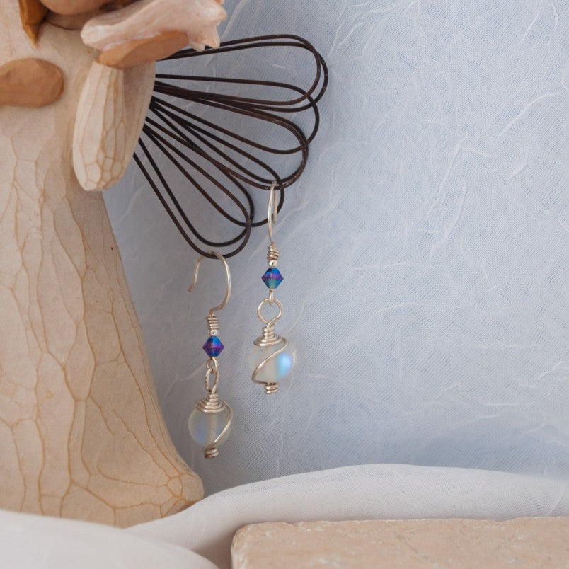 Handmade mermaid glass earrings with cosmic wire wrap and purple Swarovski bicone displayed on Willow Tree angel figurine against blue-gray background