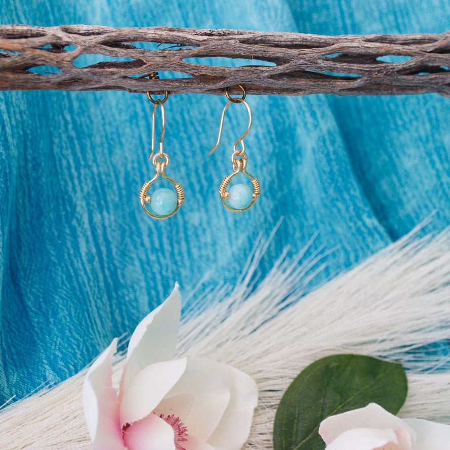 Amazonite earrings with brass wire halo and gold seed bead accents hanging from driftwood with teal water background and white magnolia flower