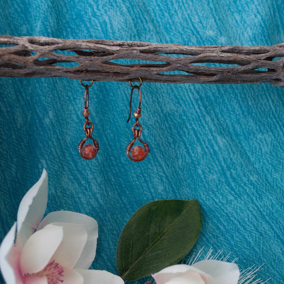Handmade Ancient Cellar agate drop earrings with antiqued copper wire halo and handcrafted hammered copper ear wires, hanging from driftwood over teal linen with white magnolia blooms — handcrafted boho gemstone earrings.