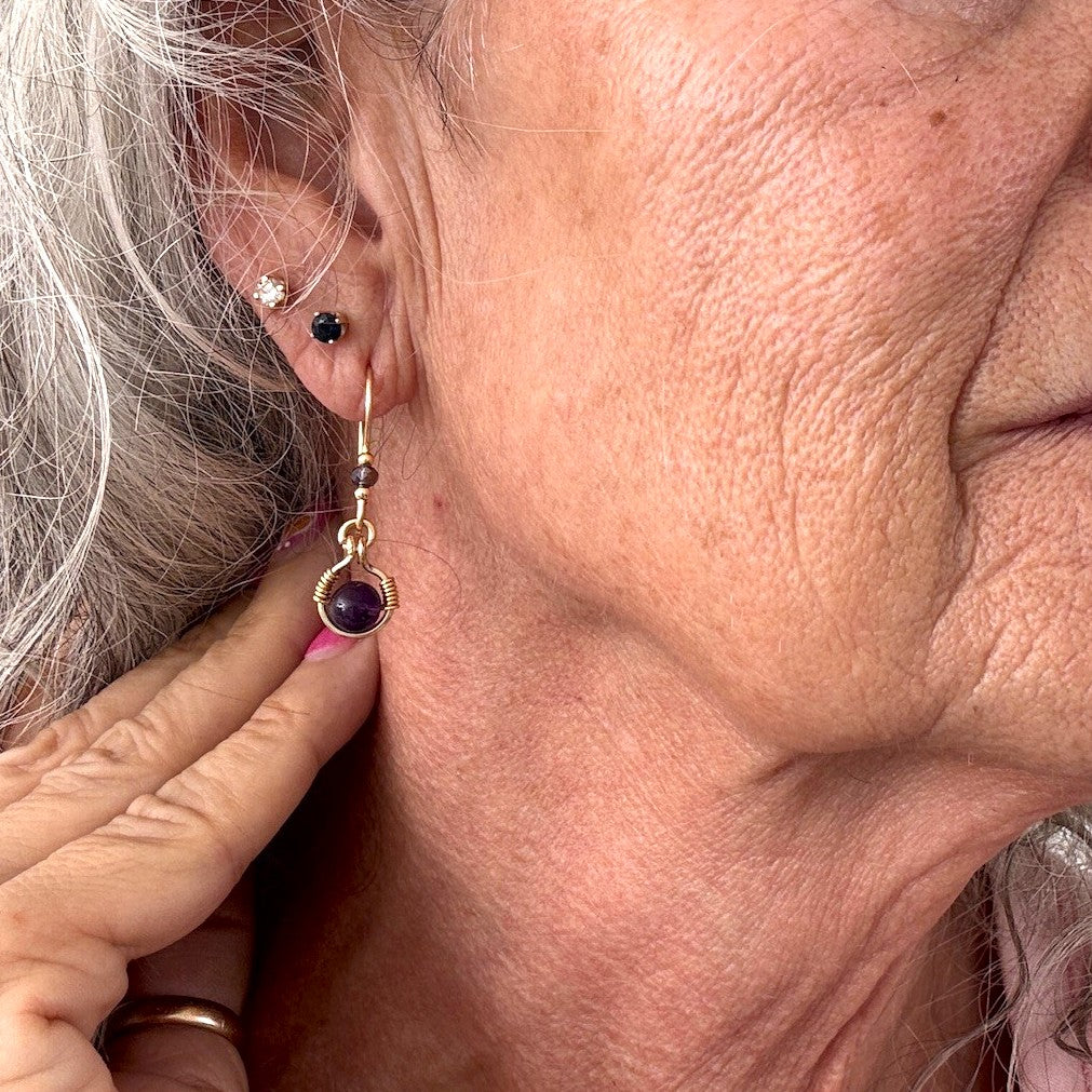 Handmade amethyst drop earrings with 6mm amethyst bead in brass wire halo and iridescent plum rondelle shown worn on model with silver hair, close-up view showing stone and construction detail