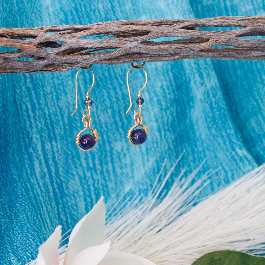 Handmade amethyst drop earrings with 6mm amethyst bead in brass wire halo, 3mm iridescent plum rondelle, gold-tone spacer beads, and handcrafted fishhook ear wires, hanging from driftwood over teal water with white magnolia flowers.