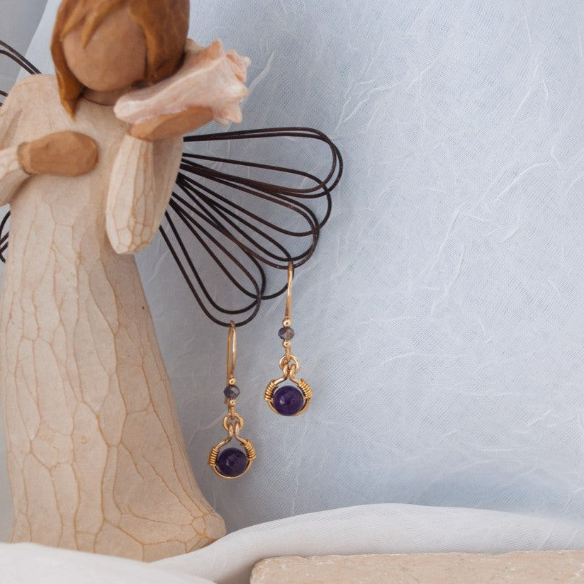 Front view of handmade amethyst drop earrings with 6mm amethyst bead in brass wire halo and handcrafted fishhook ear wires, displayed on a Willow Tree angel figurine against a blue-gray backdrop.