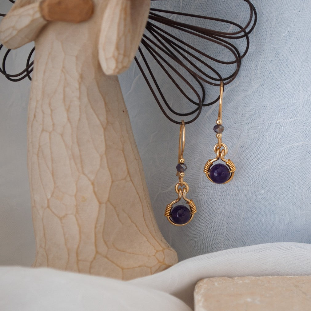 Front view of handmade amethyst drop earrings with 6mm amethyst bead in brass wire halo, 3mm iridescent plum rondelle, and handcrafted fishhook ear wires, displayed on a Willow Tree angel figurine.