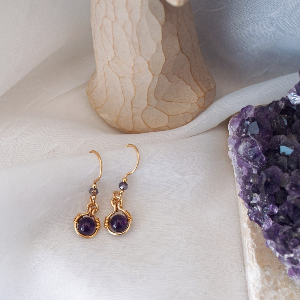 Handmade amethyst drop earrings with 6mm amethyst bead in brass wire halo and 3mm iridescent plum rondelle, laid flat beside a Willow Tree angel figurine and raw amethyst crystal cluster.