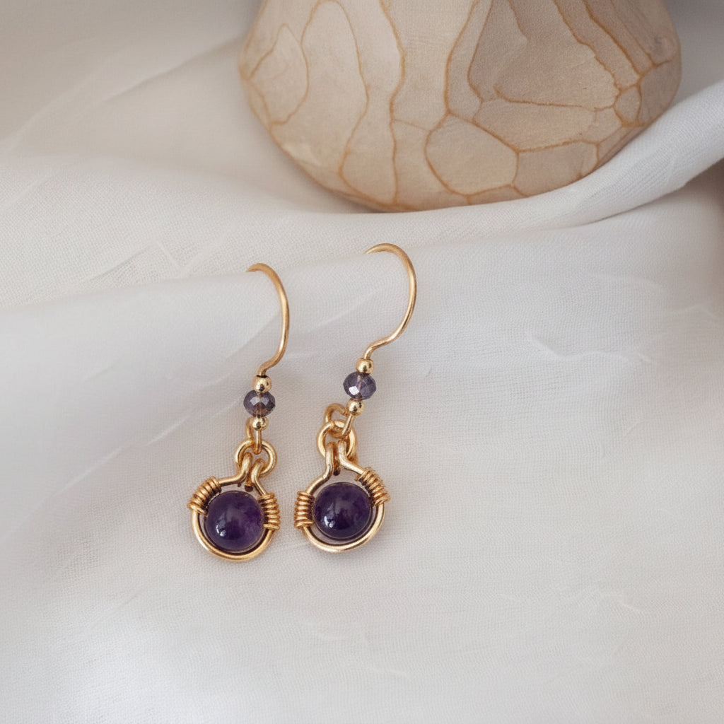 Close-up of handmade amethyst drop earrings with 6mm amethyst bead in brass wire halo, 3mm iridescent plum rondelle, and gold-tone spacer beads, laid on a white surface beside a Willow Tree angel figurine.