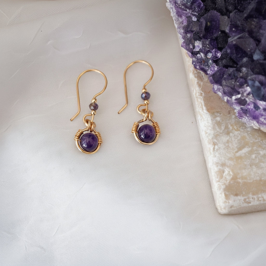 Handmade amethyst drop earrings with 6mm amethyst bead in brass wire halo and 3mm iridescent plum rondelle, laid on a white surface beside a raw amethyst crystal cluster.