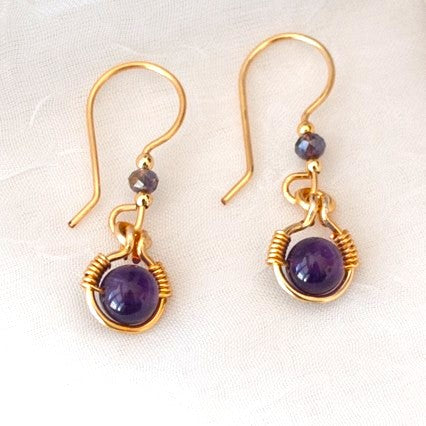 Close-up of handmade amethyst drop earrings with 6mm amethyst bead in brass wire halo, 3mm iridescent plum rondelle with gold-tone spacers, and handcrafted gold-tone fishhook ear wires, displayed on a white linen background.
