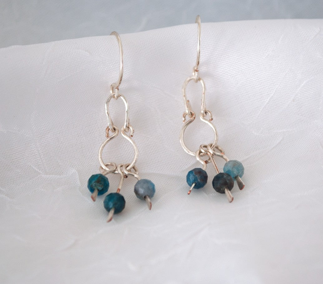 Handmade apatite earrings with hammered silver-plated oval frame, round frame, and three paddle dangles each holding a 4mm faceted apatite bead in teal and slate, displayed on white linen background