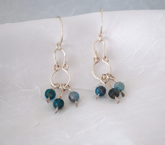 Handmade apatite earrings with hammered silver-plated oval frame, round frame, and three paddle dangles each holding a 4mm faceted apatite bead in teal and slate, displayed on white linen background