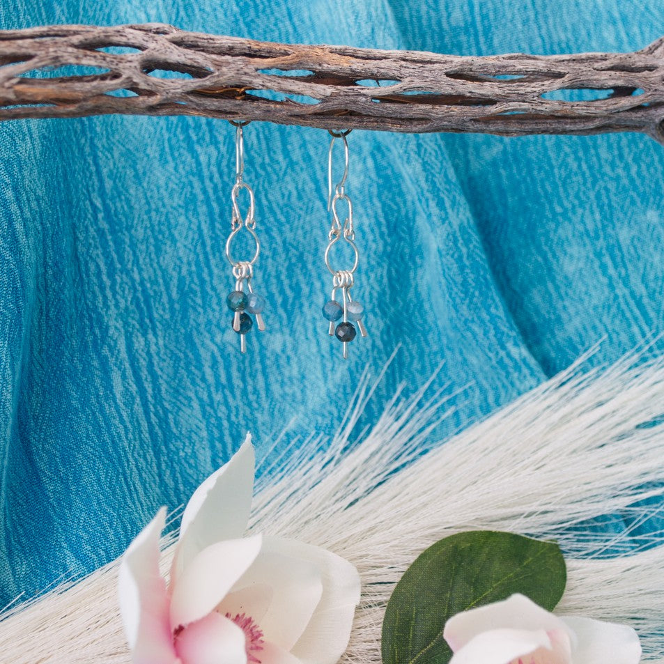 Handmade apatite chandelier earrings with hammered silver frames and three teal apatite bead dangles hanging from driftwood with teal water background and white magnolia flower