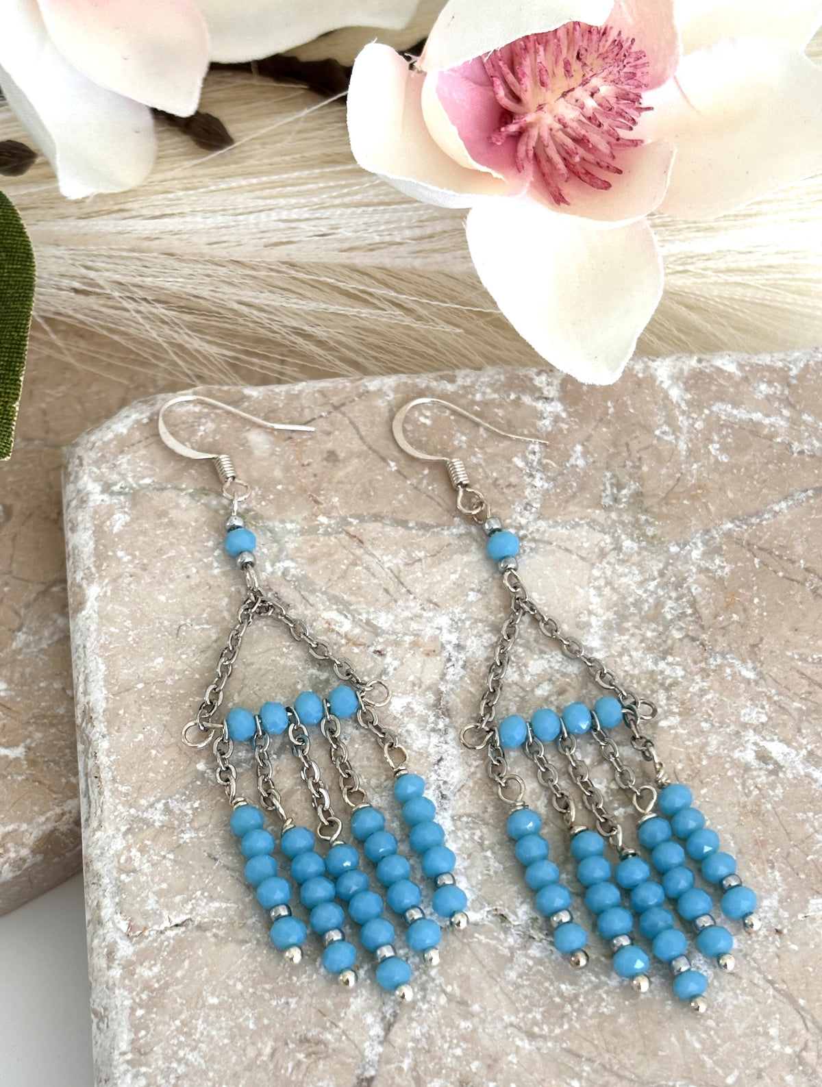 Pair of turquoise color chandelier earrings with faceted rondelle beads and silver-tone chain, displayed on travertine tile with magnolia flower