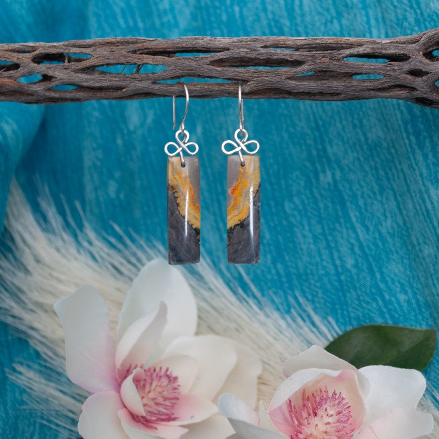 Bumble bee jasper earrings with silver-tone handcrafted bail and ear wires hanging from driftwood against teal linen background with magnolia flowers