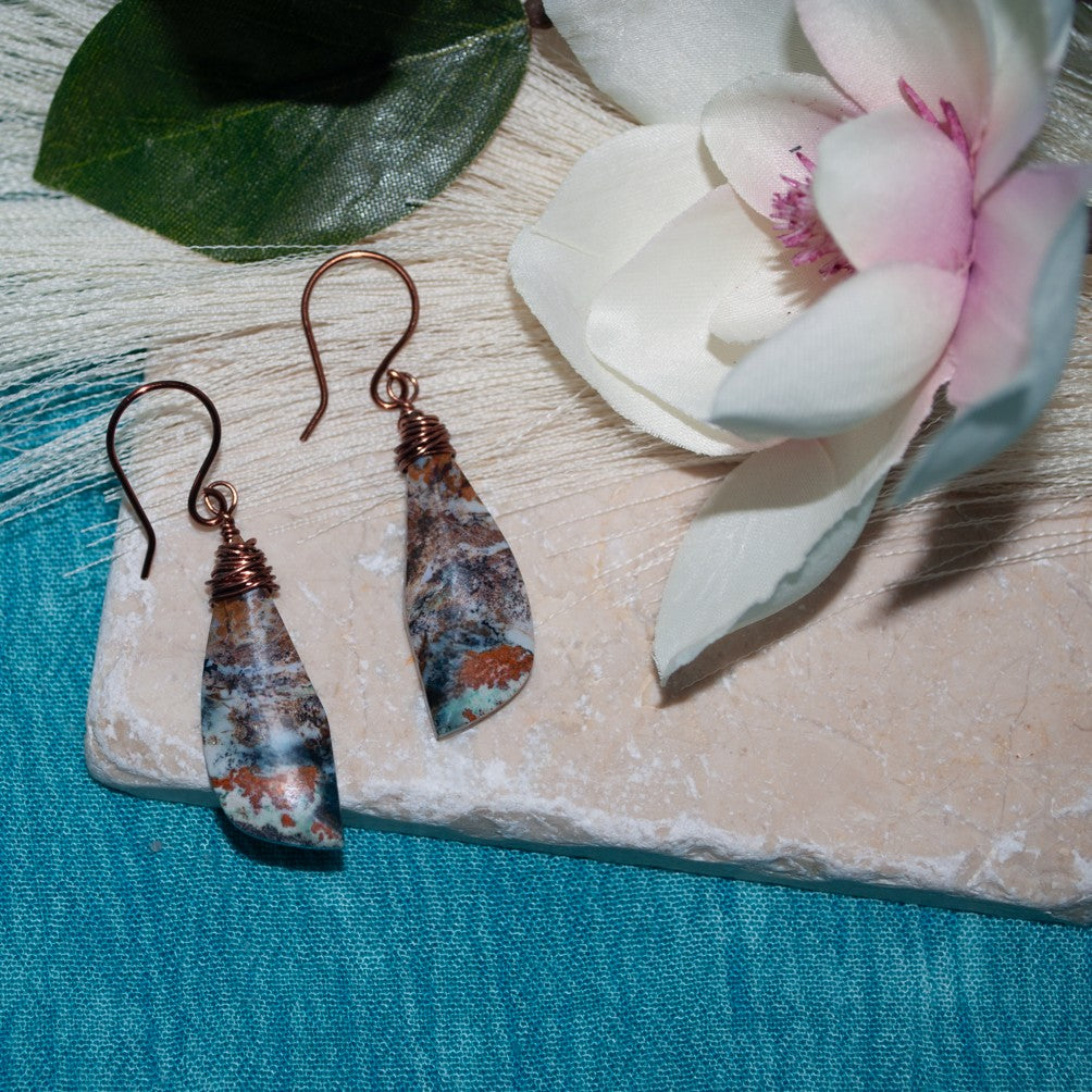 Wire-wrapped ocean jasper earrings with copper wire and handcrafted ear wires, displayed on travertine tile with magnolia flower and teal linen background