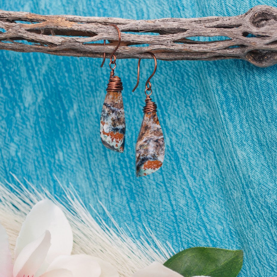 Handmade wire-wrapped ocean jasper earrings with copper wire and handcrafted copper ear wires, hanging from driftwood against a teal linen background