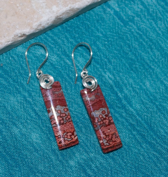 Close view of brecciated jasper earrings showing terracotta and gray stone patterning with handcrafted silver-tone swirl bail, on teal linen surface