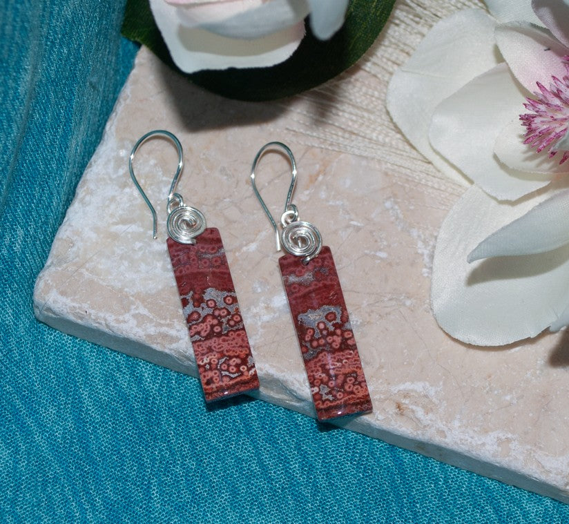 Pair of brecciated jasper earrings with silver-tone swirl bail on travertine surface with magnolia petals and teal linen backdrop