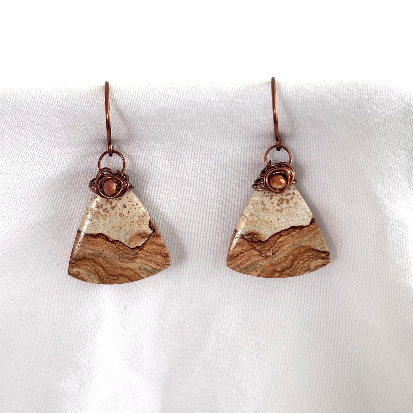 Handmade petrified wood fan-shaped drop earrings with iridescent orange crystal bead and antique copper wire wrap bail, laid on draped white fabric showing natural wood grain banding detail.