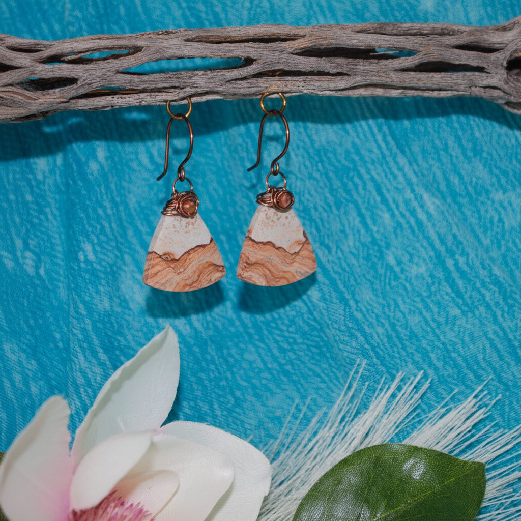Handmade petrified wood fan-shaped drop earrings with iridescent orange crystal bead and antique copper wire wrap bail, hanging from driftwood over turquoise water with a white magnolia flower.