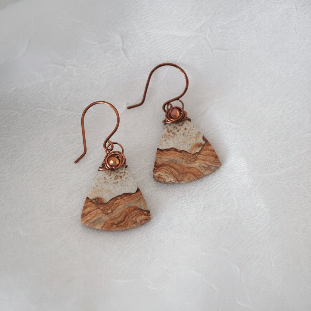 Handmade petrified wood fan-shaped drop earrings with iridescent orange crystal bead and antique copper wire wrap bail, laid on white fabric.