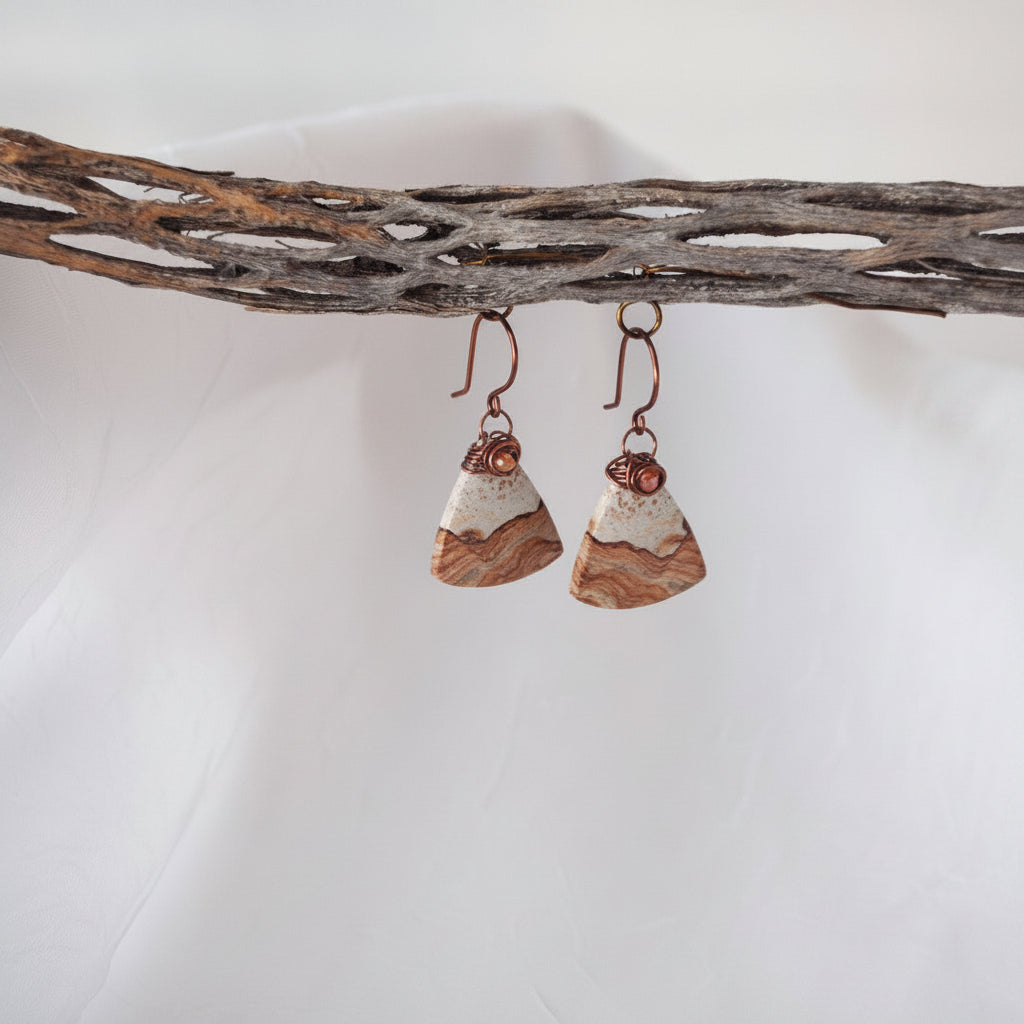 Handmade petrified wood fan-shaped drop earrings with iridescent orange crystal bead and antique copper wire wrap bail, hanging from a rustic driftwood branch.