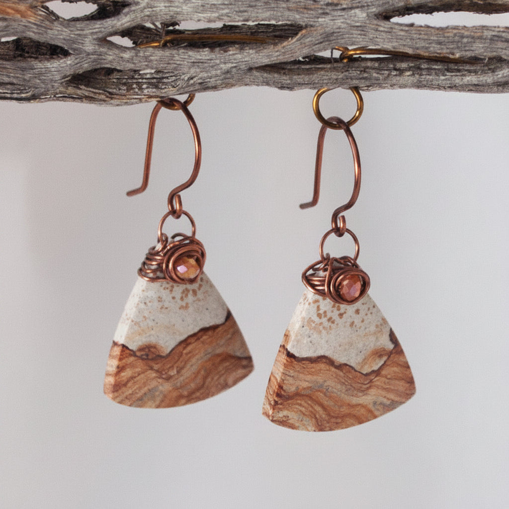 Close-up detail of handmade petrified wood fan-shaped drop earrings showing iridescent orange crystal bead and antique copper wire wrap bail, hanging from a rustic driftwood branch.