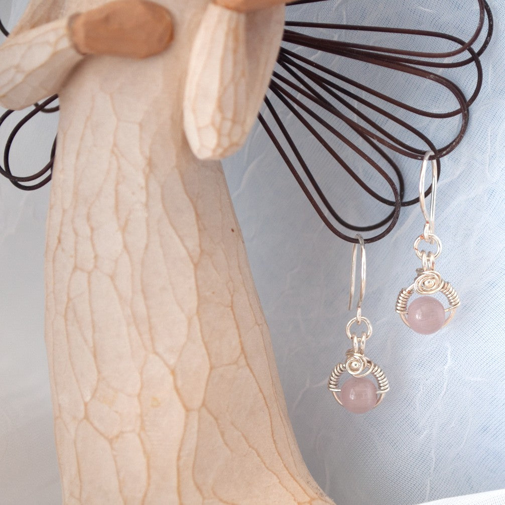 Handmade pink cat's eye earrings with silver-plated wire halo and wire-wrapped rose displayed on Willow Tree angel figurine against blue-gray background
